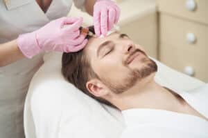 Young man smiling while cosmetologist making a Botox injection into his forehead.