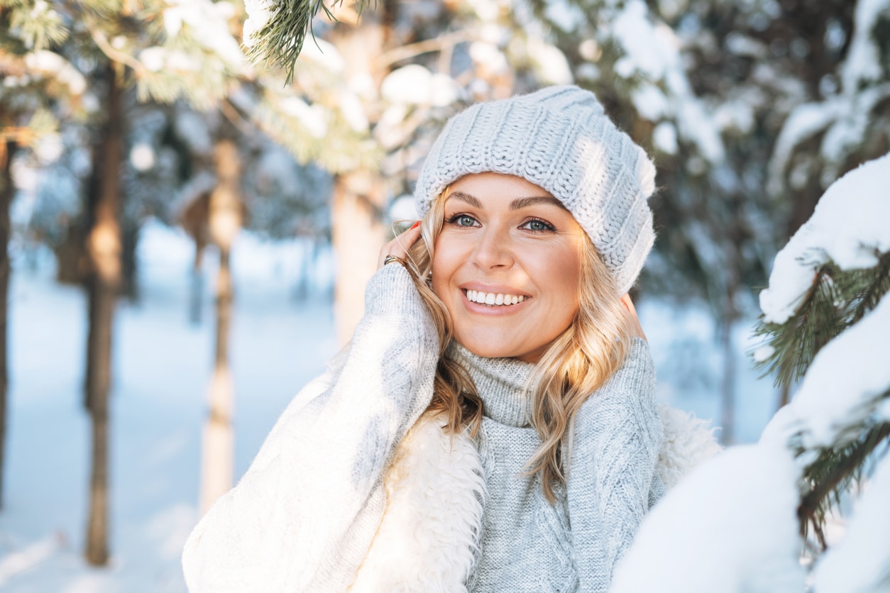 A beautiful woman in a white winter outfit stands in a snowy, tree-filled area but her complexion is protected by the best skin care.