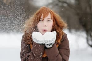 A warmly dressed woman with beautiful skin stands outside and blows a handful of snow towards the camera.