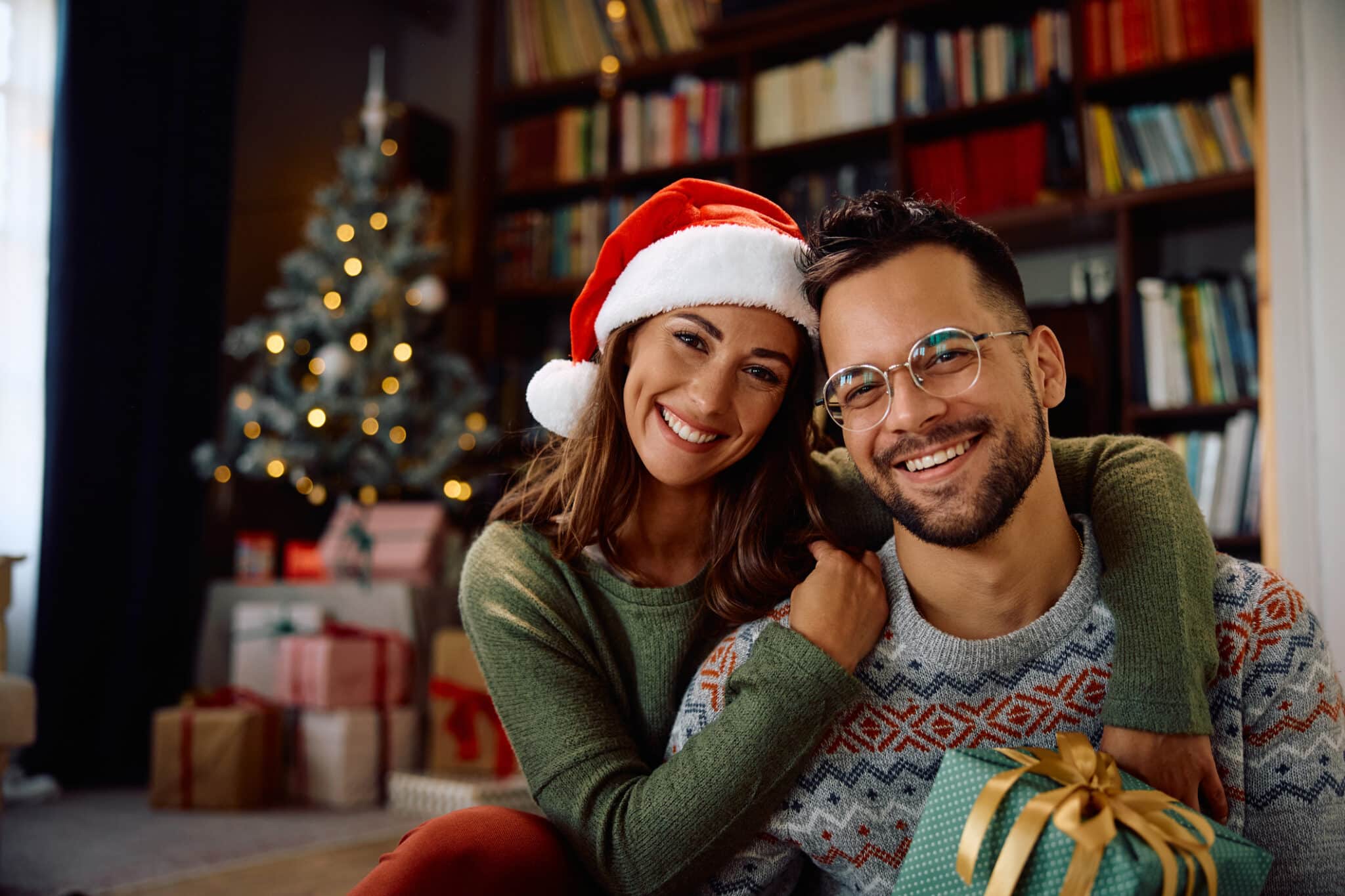 A couple sits in their holiday-decorated living room, smiling at the camera because they are proud of their Christmas gift ideas like a spa membership and vision care plan!