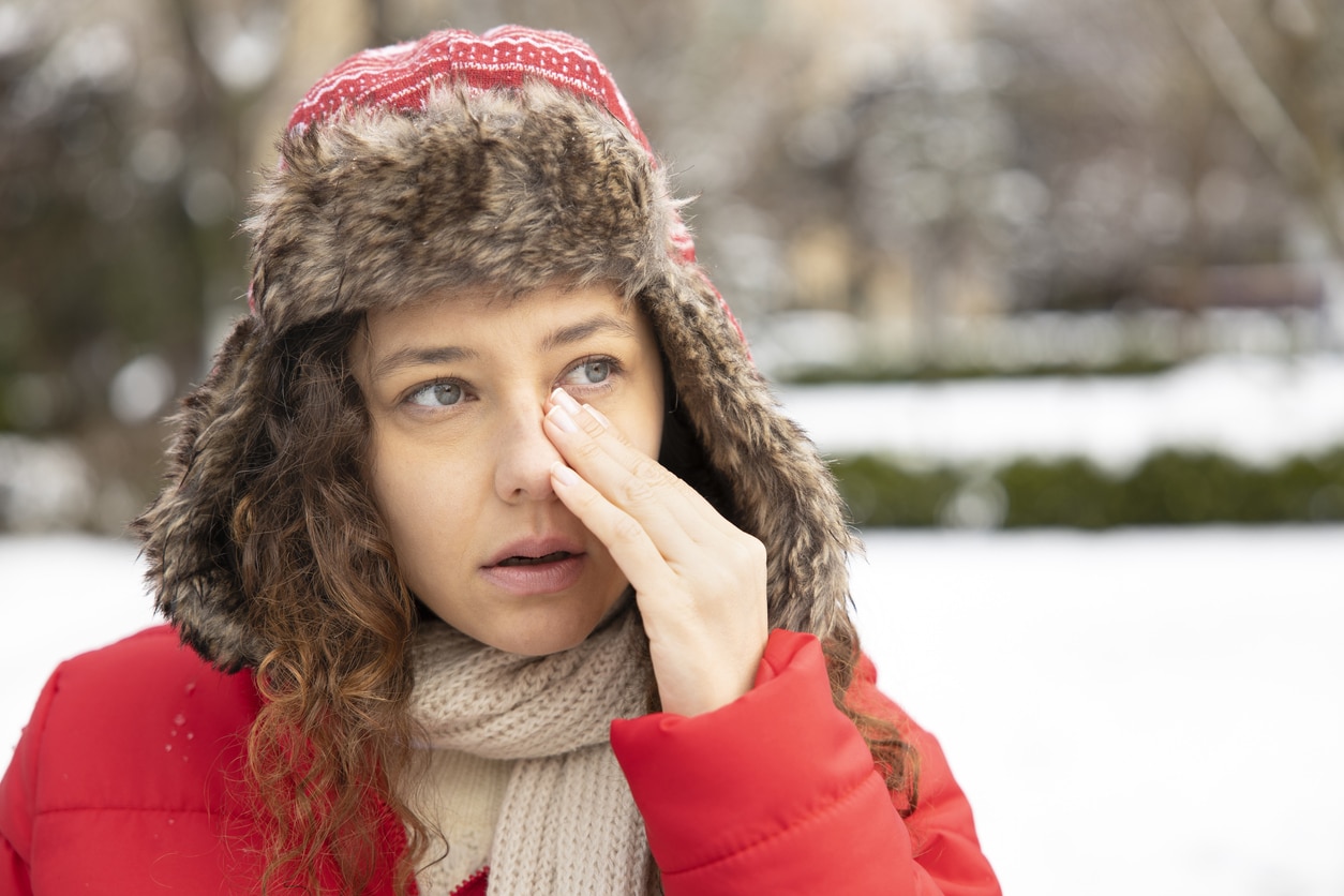 A woman dressed in warm winter clothing stands outdoors in a snowy landscape, rubbing her dry eyes that are irritated by the cold weather.