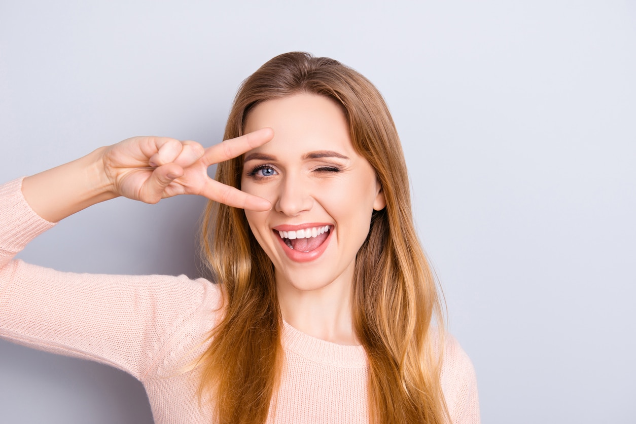 A young woman winks and smiles, holding up a sideways peace sign to frame her eye and eyebrow, showing off the results of beautiful brow and lash treatments.