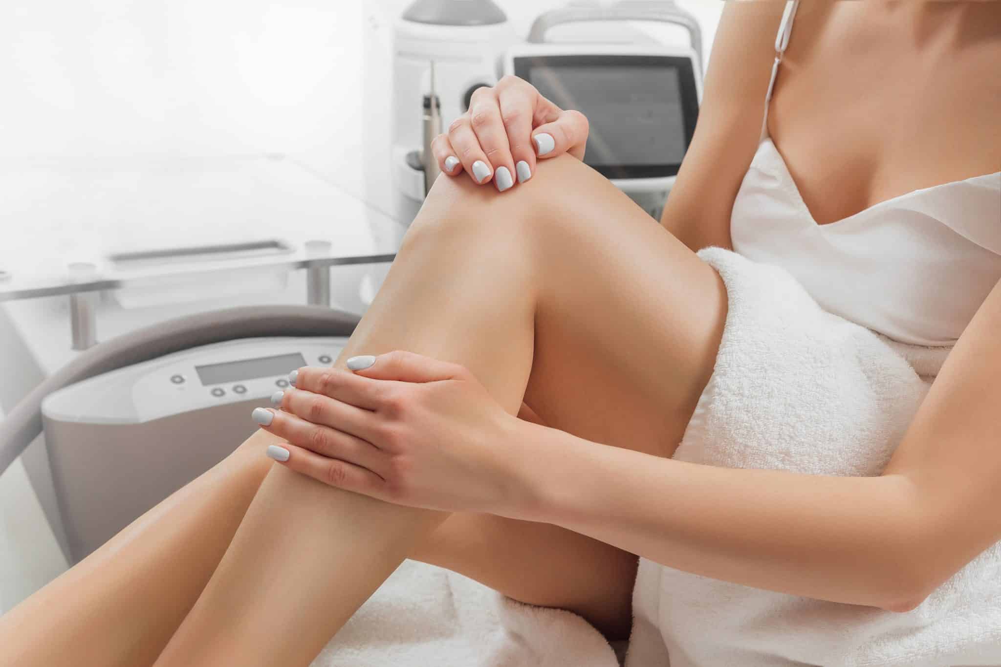 A woman sitting on a spa treatment table runs her hand over the bare skin of her leg, enjoying the benefits of laser hair removal.