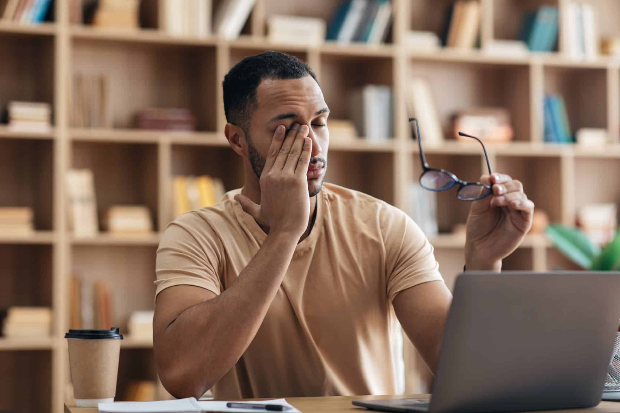 A man sits at a laptop, holding a pair of glasses as he rubs his eyes with one hand, experiencing vision fatigue and the effects of computer vision syndrome.