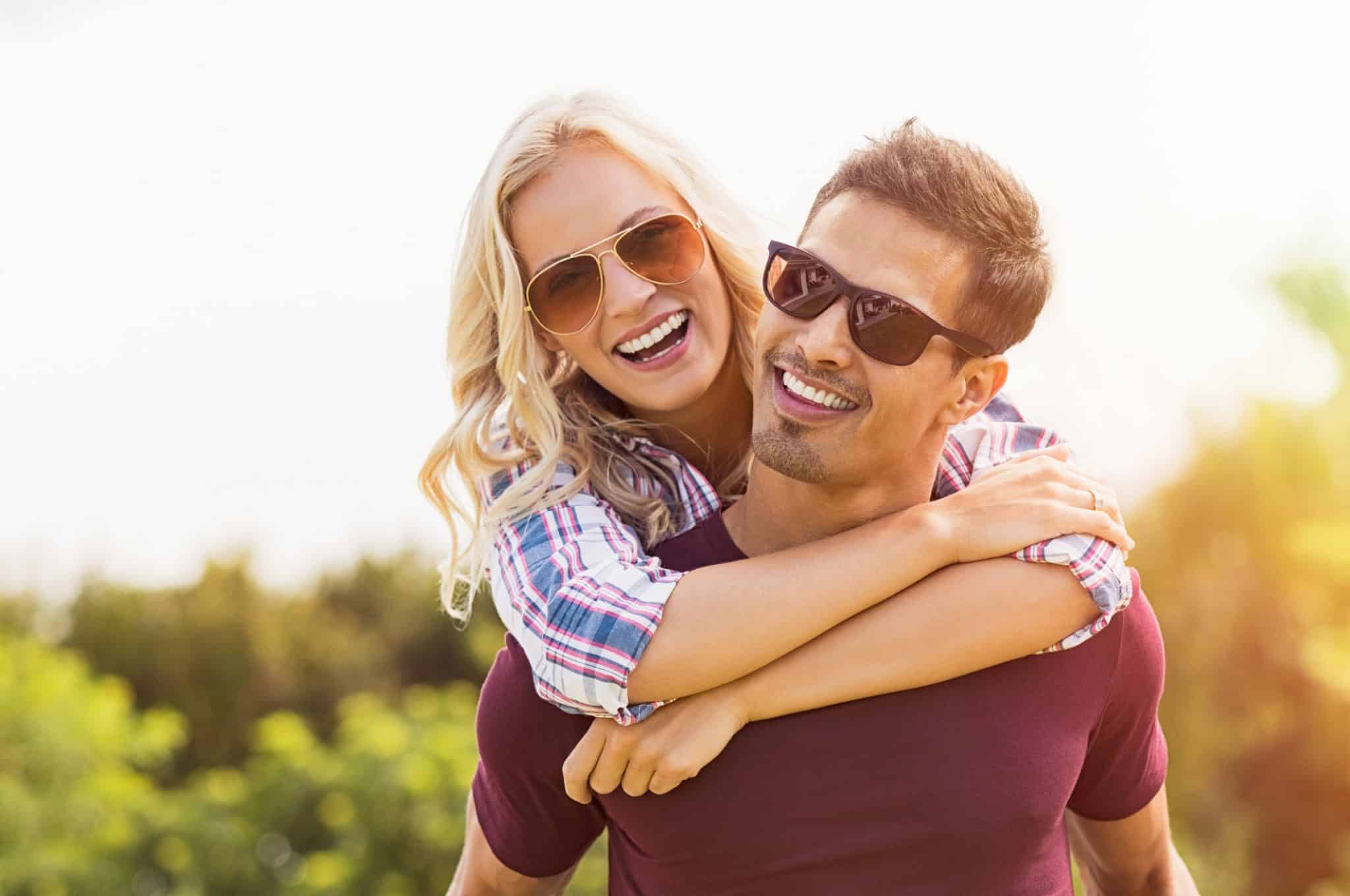 Closeup of a young man carrying a beautiful woman on his back. They both wear sunglasses to protect their eyes from UV damage as they enjoy the summer day.