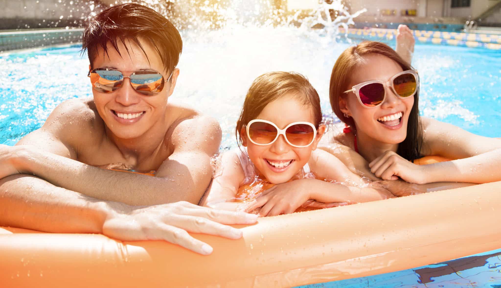 A happy family plays together in a swimming pool, wearing sunglasses and confident about their understanding of summer eye health for swimming.