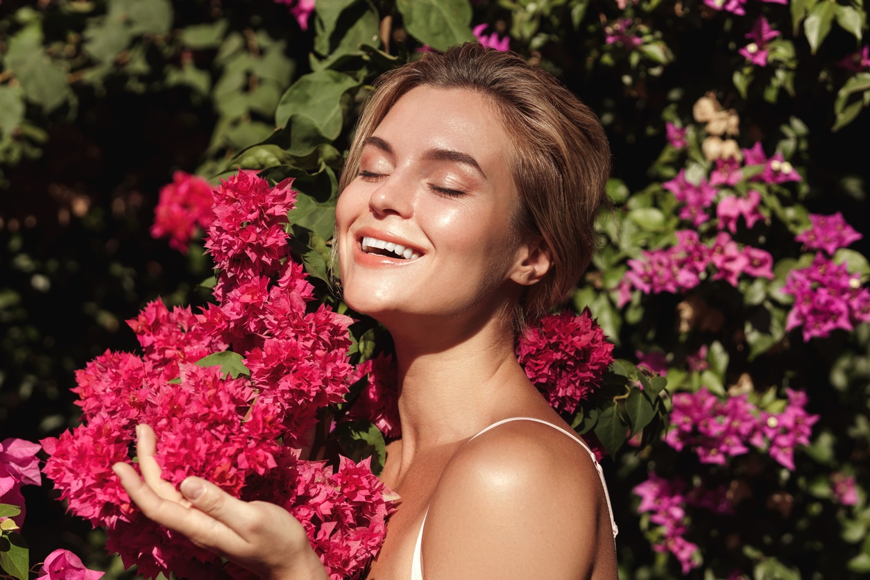 A woman with smooth, glowing skin poses outdoors near pink flowers, reflecting fresh spring skincare results.