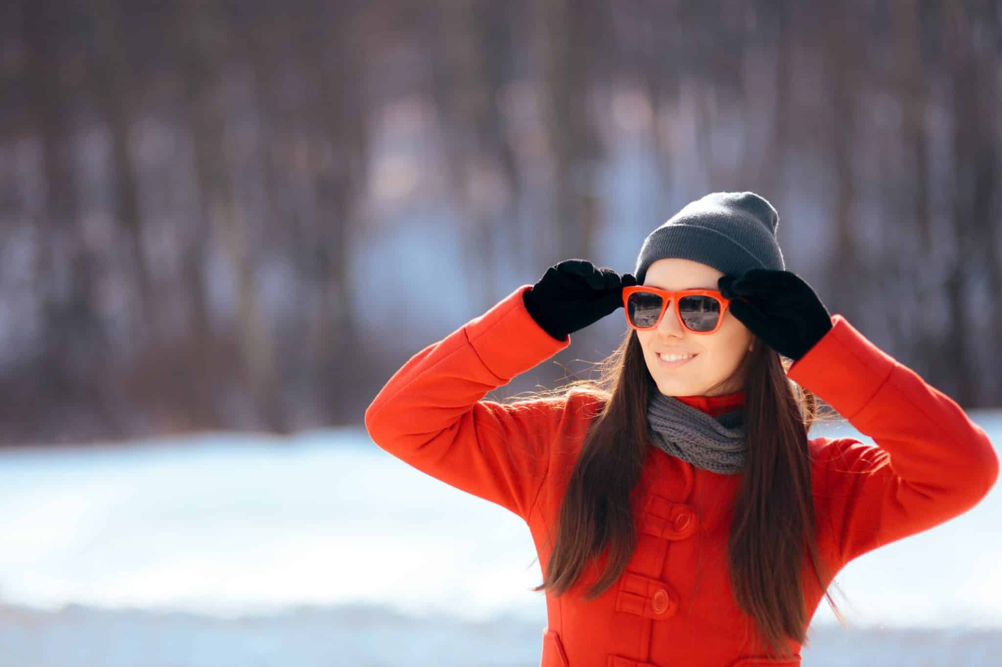 A woman with beautiful skin wears sunglasses to protect her eyes from UV rays bouncing off of the snow.